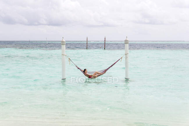 Femme en maillot de bain rouge couché dans hamac balançant sur la ligne de surf océanique relaxant aux Maldives par temps nuageux — Photo de stock