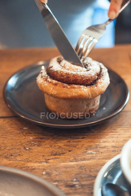 High angle of unrecognizable person cutting sweet cinnamon bun with knife at wooden table in cafe — Stock Photo