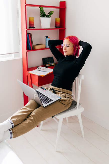 Freelancer jovem focado em roupas casuais sentado na cadeira com olhos fechados usando laptop enquanto trabalhava no projeto em apartamento moderno claro — Fotografia de Stock