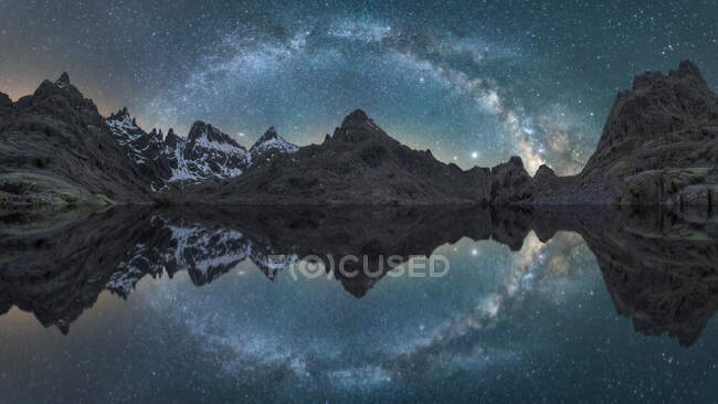 Impresionante paisaje nocturno de ásperas montañas rocosas con nieve cerca de un lago tranquilo con superficie de agua lisa que refleja el cielo con la brillante Vía Láctea - foto de stock