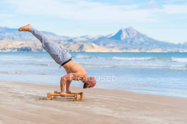 Side view full length of male athlete performing handstand with parallel bars on sandy coast with ocean waves on background — Stock Photo