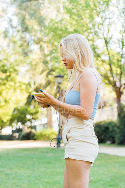 Vista lateral de una mujer tranquila con ropa de verano escuchando música en los auriculares mientras está de pie en el parque - foto de stock