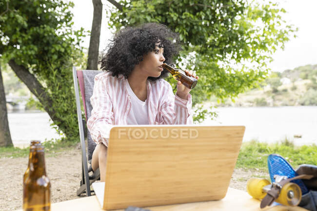 Молодая латиноамериканка с вьющимися волосами потягивает пиво из бутылки и смотрит в сторону, сидя за столом с ноутбуком в выходные дни в сельской местности — стоковое фото