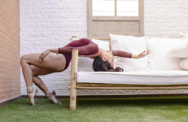 Вид збоку на гнучку витончену балерину у взутті та боді-костюмі, що балансує на топах та показує бік, спираючись на диван на терасі — стокове фото