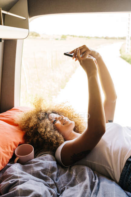 Donna anonima con acconciatura Afro sdraiata a testa in giù sul letto in roulotte e navigando sul cellulare nella soleggiata giornata estiva — Foto stock