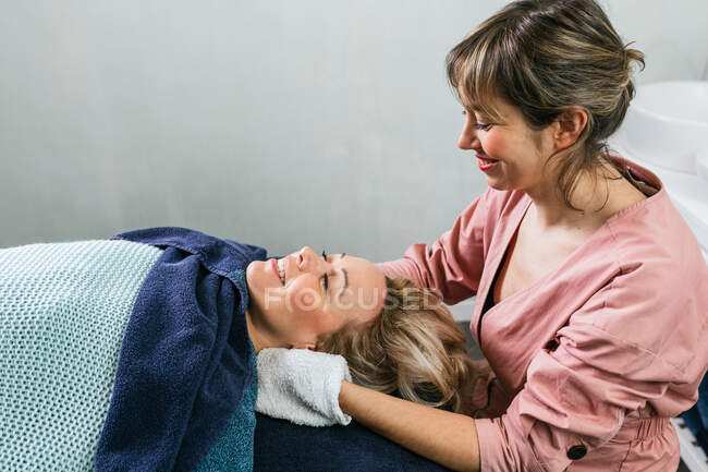Vue latérale de l'esthéticienne gaie essuyant le visage de la cliente avec une serviette pendant la routine de soins de la peau dans le salon de spa — Photo de stock