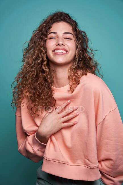 Mujer sonriente con el pelo rizado usando sudadera de pie mirando a la cámara en el estudio con fondo azul - foto de stock
