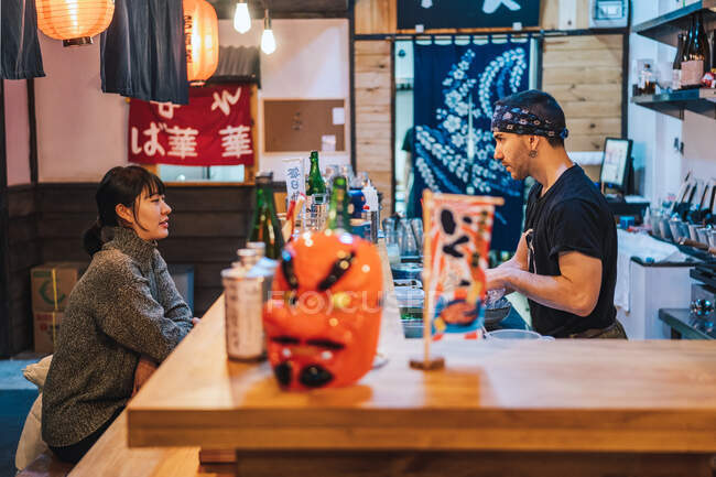 Vista laterale della donna asiatica in abbigliamento casual seduta al bancone e che parla con il lavoratore maschile del moderno ramen bar — Foto stock