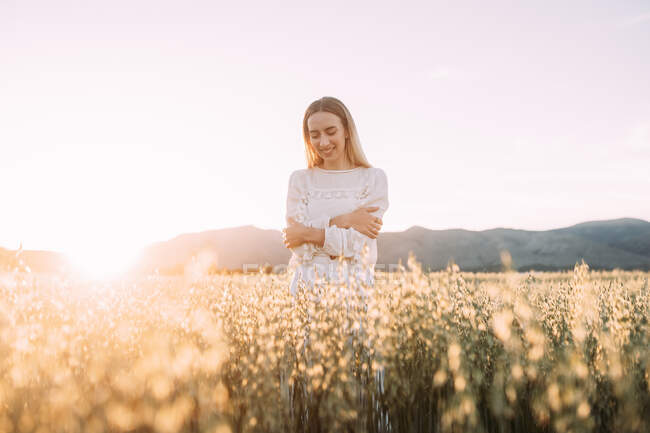 Cheerful blond female in light white dress embracing arms while standing in high blooming grass in field against sunset light — Stock Photo