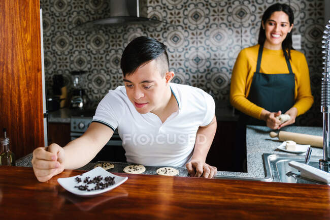 Deleitado adolescente étnico con síndrome de Down decoración de galletas con chips de chocolate mientras se prepara la pastelería con la madre sonriente en casa - foto de stock