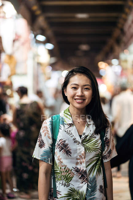 Happy Asian female traveler in tropical shirt with backpack smiling at camera while standing on bazaar against blurred crowd in Doha — Stock Photo