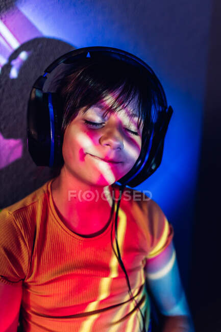 Chica alegre en ropa casual tableta de navegación y escuchar canciones favoritas con los ojos cerrados a través de auriculares mientras está sentado en luces de neón - foto de stock
