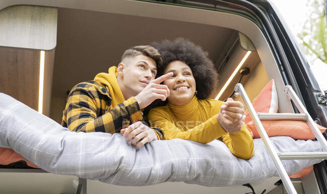 Low angle of African American woman smiling with young boyfriend while lying down pointing away on bed in van during road trip looking away — Stock Photo