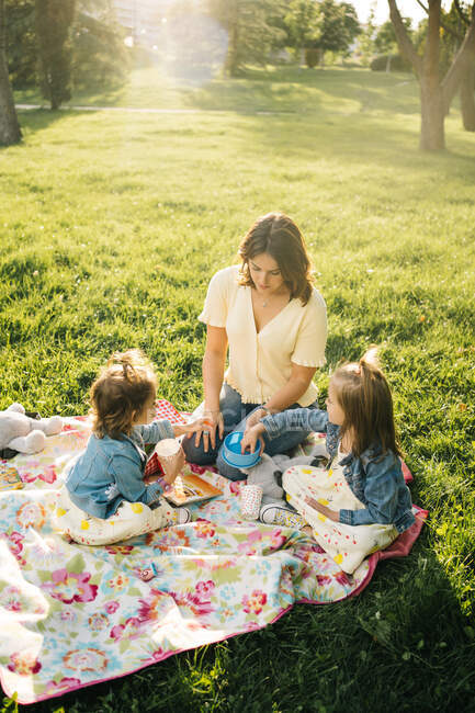 Mujer joven feliz con hijas pequeñas disfrutando de un picnic en el prado verde mientras pasan el día de verano juntos en el parque - foto de stock