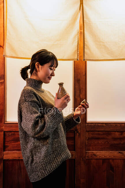 Vista laterale della felice donna asiatica in maglione in piedi vicino alla porta con bevanda tradizionale nel caffè — Foto stock