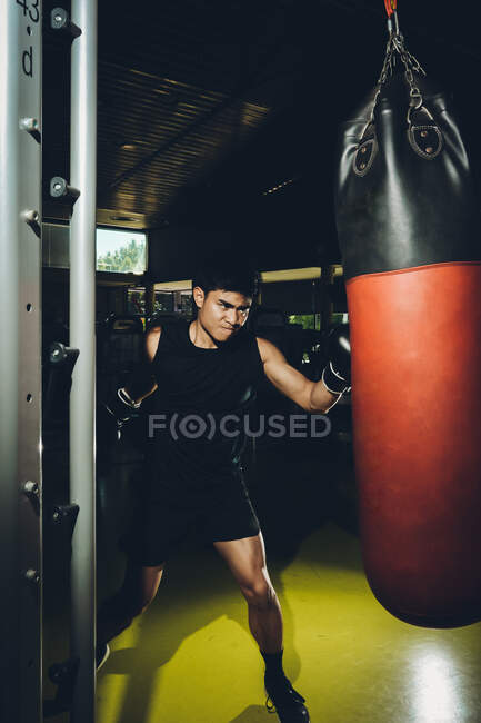 Junger fokussierter asiatischer Mann beim Boxtraining, während er mit schwerem Boxsack in einem modernen Fitnessstudio trainiert — Stockfoto