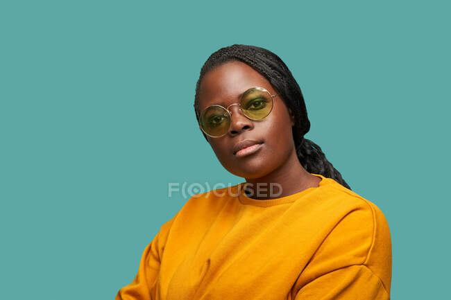 Mujer afroamericana regordeta sin emociones en suéter amarillo y gafas de sol de pie mirando a la cámara contra la pared azul - foto de stock