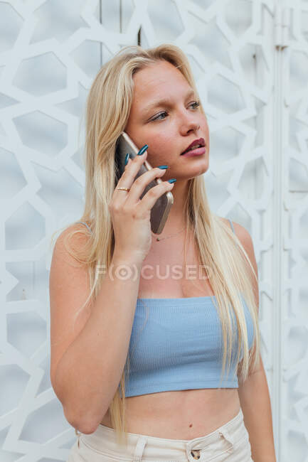 Mujer tranquila con el pelo rubio y en traje de verano de pie en la ciudad y hablando por teléfono móvil mientras mira hacia otro lado - foto de stock