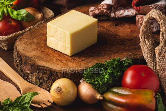 Блок сыра на деревянной стойке рядом с сырым луком и тканый мешок против органических лопаток и листьев базилика — стоковое фото