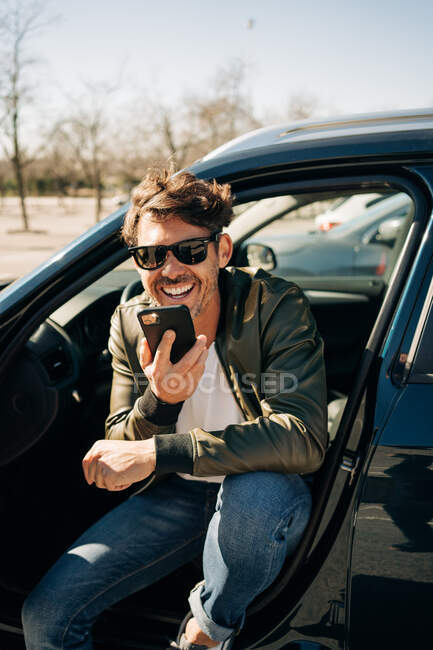 Happy male in sunglasses sending voice message on cellphone while sitting in automobile on sunny day — Stock Photo