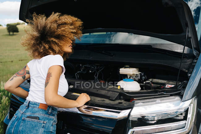 Back view of unrecognizable traveling female looking under opened hood of camper van while having problems during trip through summer nature — Stock Photo