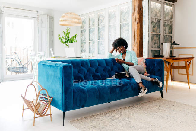Schöne junge Afroamerikanerin in lässiger Kleidung surft mit ihrem Smartphone in die Kamera, während sie sich zu Hause auf dem gemütlichen blauen Sofa ausruht — Stockfoto