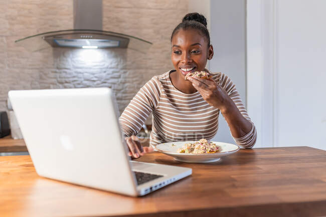 Афроамериканська самиця насолоджується смачним патчем з верхівкою, а вдома переглядає в Інтернеті нетбук на кухні. — стокове фото