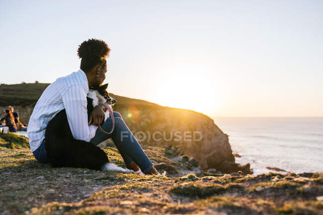 Side view of young African American female owner with Border Collie dog spending time together on beach near waving sea at sunset contemplating views — Stock Photo