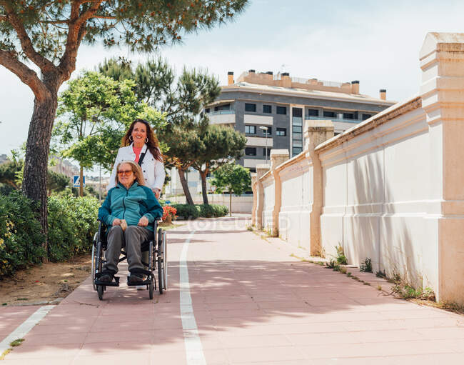 Heureuse femme adulte regardant la caméra tout en poussant le fauteuil roulant avec la mère aînée pendant la promenade dans la rue en ville en été — Photo de stock