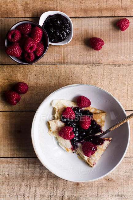 Vista dall'alto di deliziose crepes con marmellata di fragole dolci posizionate sul piatto vicino al cucchiaio sul tavolo di legno — Foto stock