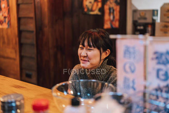 Asian woman in casual wear sitting at wooden counter while waiting for order in ramen bar — Stock Photo