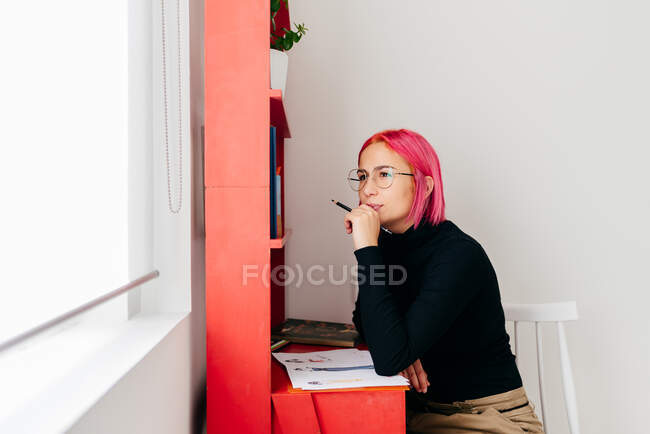 Молода вдумлива креативна жінка-дизайнерка з рожевим волоссям у повсякденному вбранні та окулярах малює ескіз під час роботи за столом вдома — стокове фото