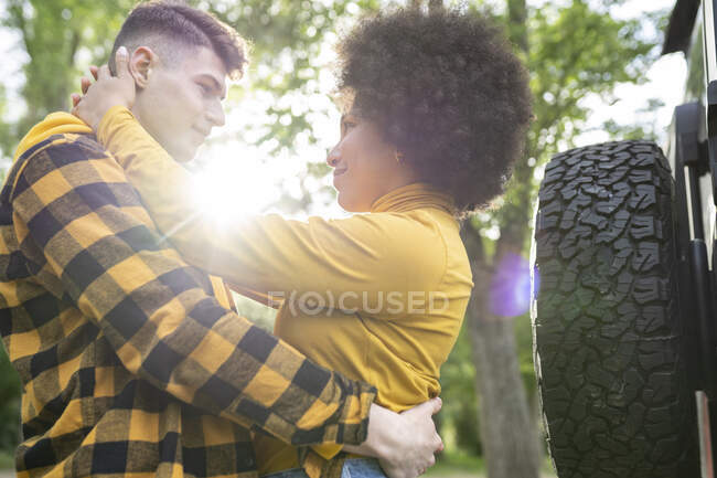 Baixo ângulo de homem e mulher multiétnicos jovens em roupas casuais abraçando uns aos outros enquanto estão perto do carro no dia ensolarado no campo — Fotografia de Stock