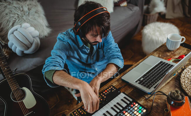De arriba joven feliz en los auriculares usando sintetizador y portátil en la mesa en casa - foto de stock