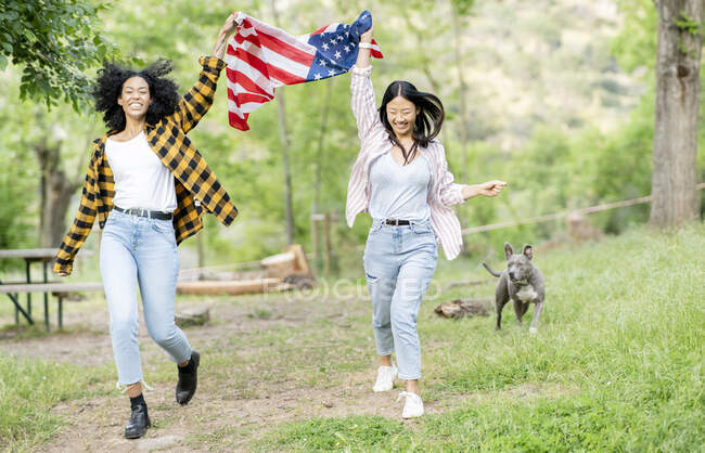 Cheerful lesbian multiracial couple of females running with national American flag along path in forest and smiling looking at camera — Stock Photo