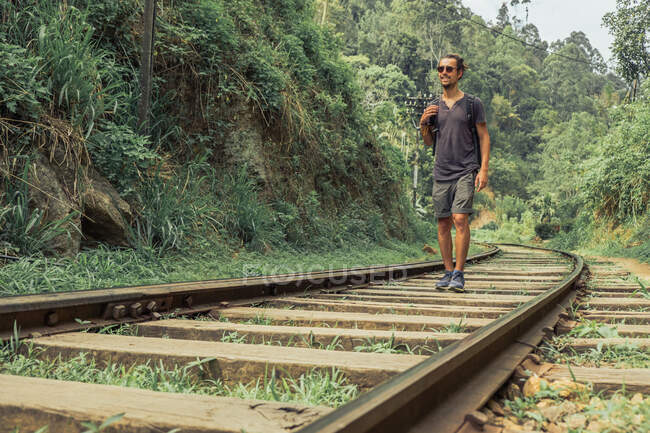 Веселий чоловік мандрівник з рюкзаком, що йде залізницею в тропічних лісах під час літніх канікул — стокове фото