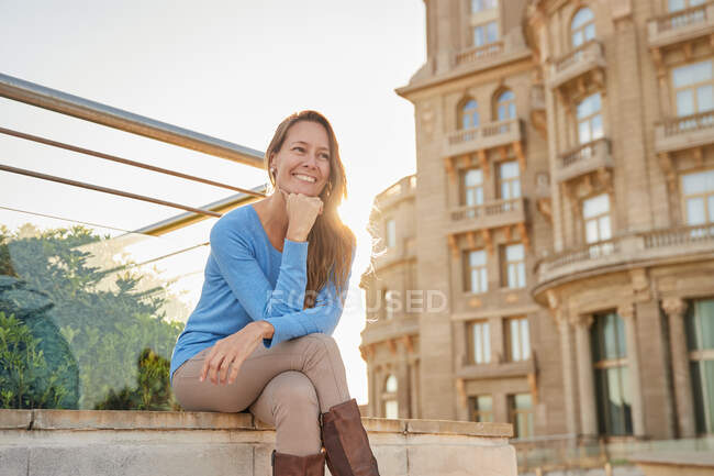 Femme adulte heureuse en tenue décontractée assise près d'une rampe avec un vieux bâtiment en arrière-plan dans la rue de la ville par une journée ensoleillée — Photo de stock