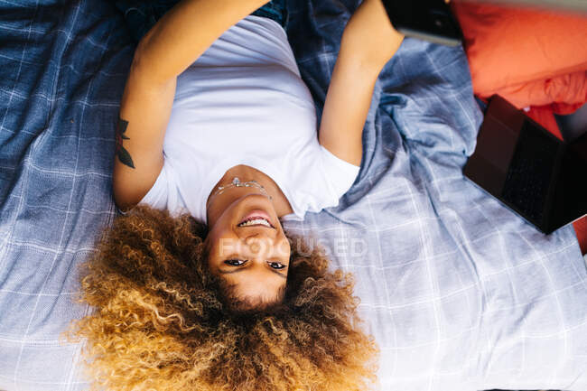 Positive black woman with Afro hairstyle lying upside up on bed in trailer and browsing mobile phone on sunny summer day — Stock Photo