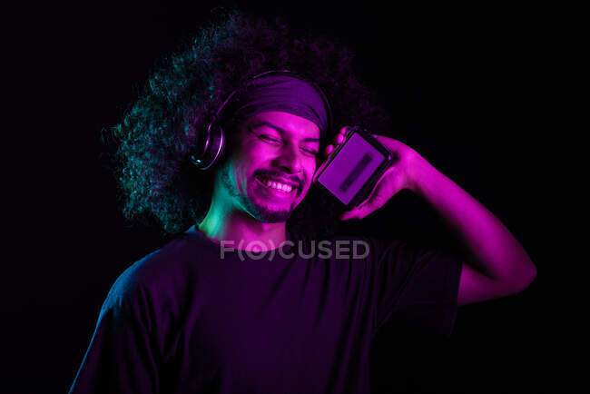 Счастливый латиноамериканец в наушниках и с ретро-кассетой, наслаждающийся песнями на черном фоне в студии с синей и фиолетовой неоновой подсветкой — стоковое фото