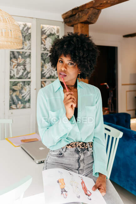 Moderna y exitosa mujer afroamericana freelancer con un atuendo elegante con cabello afro mirando la cámara mientras está sentada en la mesa y leyendo el documento en casa - foto de stock