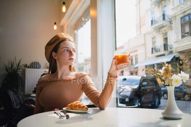 Французька самиця в береті сидить за столом у кафе з ароматичним склом кави і свіжим печеним круасаном. — стокове фото