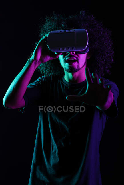 Excited Latin male with Afro hairstyle and in VR goggles experiencing virtual reality on black background in studio with neon lights — Stock Photo