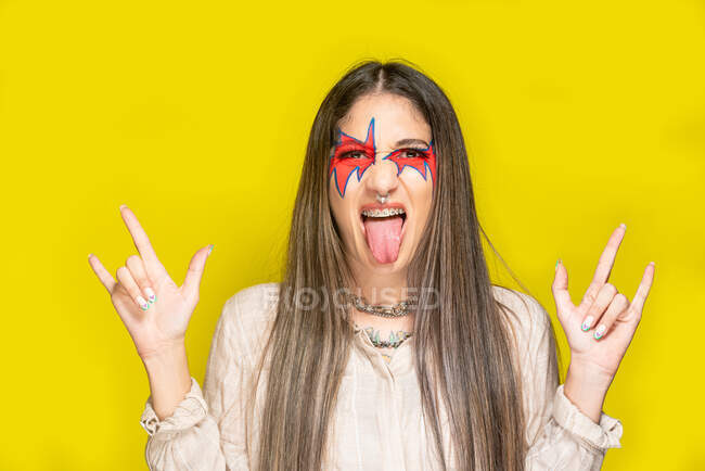 Jovem fêmea elegante com maquiagem criativa gesticulando chifres e mostrando língua contra fundo amarelo — Fotografia de Stock