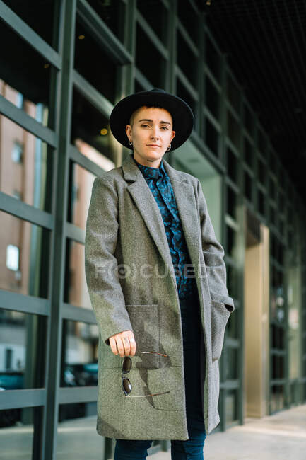 Молодой трансгендер в шикарном пальто и шляпе смотрит в камеру при свете дня — стоковое фото