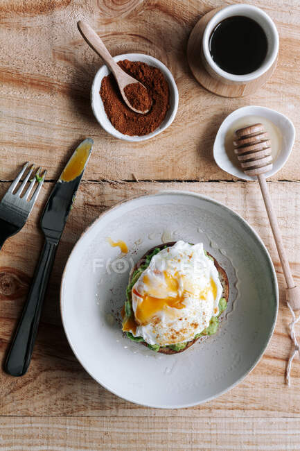 Apetitivo trozo de pan de grano con aguacate y huevo escalfado en la parte superior colocado en el plato en la mesa de madera - foto de stock