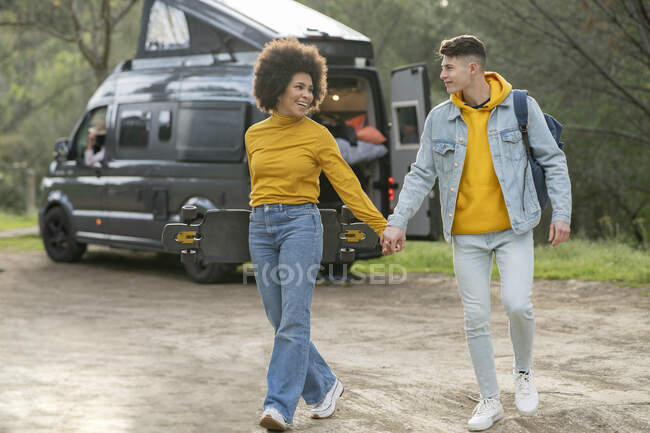 Diverse young man and woman with longboard holding hands and walking on countryside road on summer weekend day near caravan — Stock Photo
