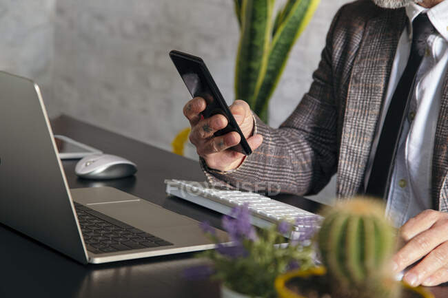 Cortar irreconhecível tatuado executivo masculino em checkered jaqueta mensagens de texto no celular contra laptop durante o teletrabalho — Fotografia de Stock