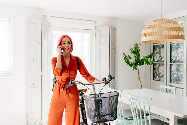 Jovem designer feminina criativa em roupas da moda e óculos falando no smartphone enquanto está de pé com bicicleta no apartamento leve moderno — Fotografia de Stock