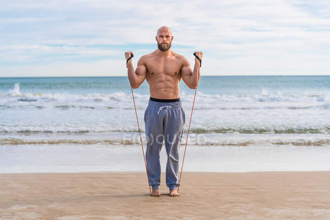 Жестокий мужчина без рубашки смотрит в камеру и делает боковые мухи с устойчивой тренировкой на берегу моря. — стоковое фото
