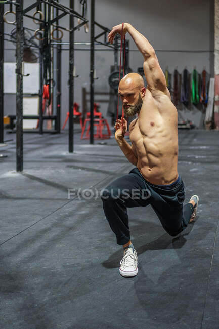 Full body shirtless bearded guy stretching elastic band with arms and lunging during functional workout in gym — Stock Photo
